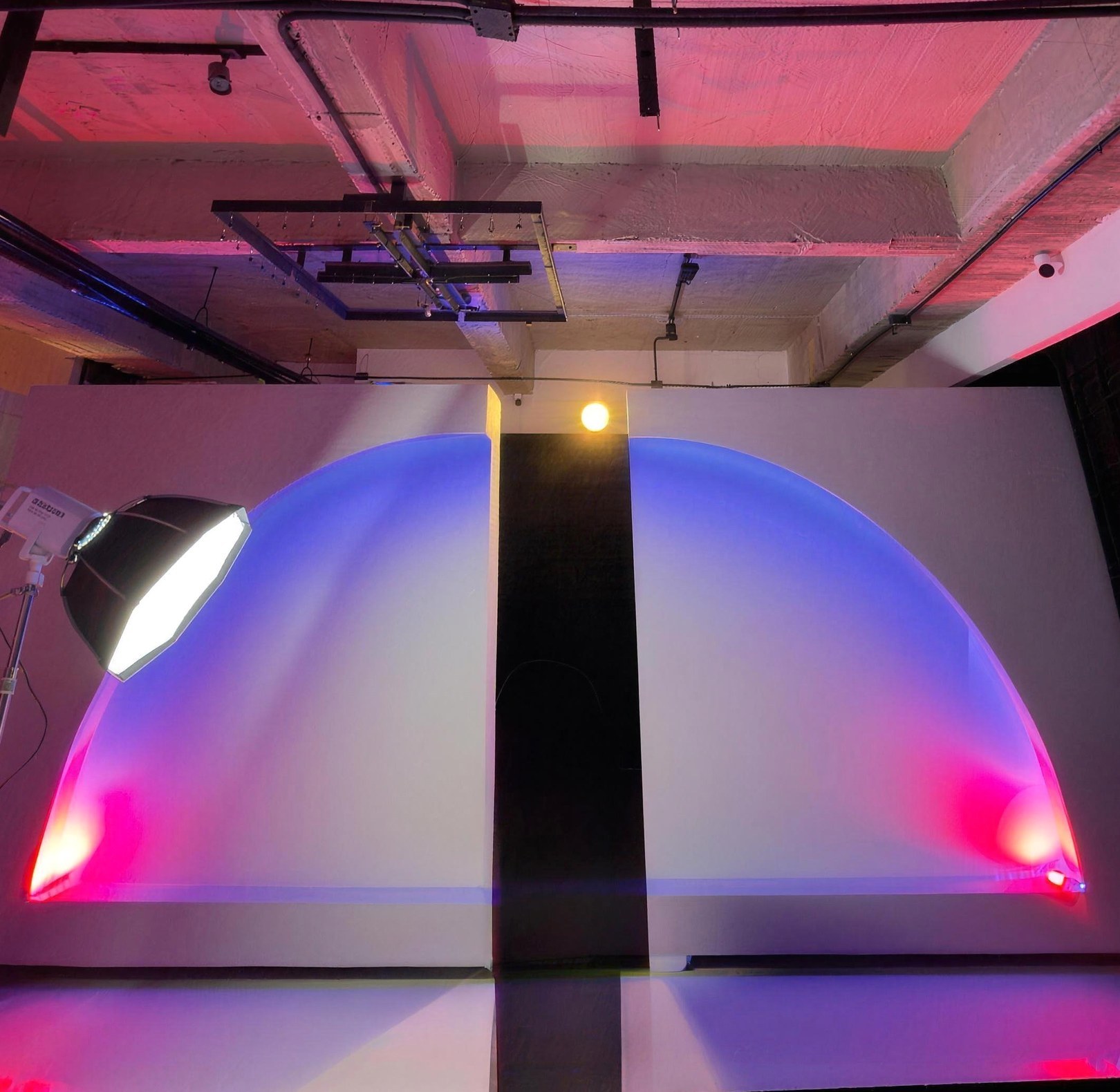 Dos arcos de luz azul y magenta proyectados sobre un muro blanco con panel negro central, con luz Amaran